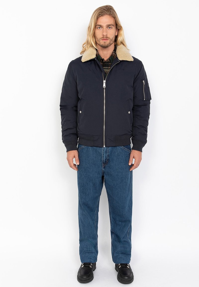 Chaqueta bomber acolchada azul marino con cuello de piel sintética, cerradura frontal, bolsillos laterales y un bolsillo con cremallera en el brazo; combinada con jeans azules y zapatos negros.