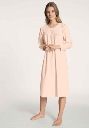 Donna in piedi a piedi nudi, che indossa un camicione lungo fino al ginocchio di un tenue rosa pallido con tessuto arricciato e delicata ricamo sul petto.