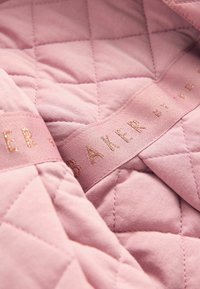 Gepolsterter pinker Stoff mit einer glatten Textur, versehen mit einem Band, das das Wort "BAKER" in glänzendem Roségold aufgedruckt hat.