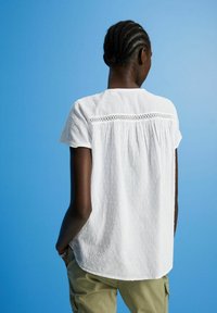 Camicia bianca a maniche corte con tessuto testurizzato e dettaglio ricamato sulla spalla, caratterizzata da una vestibilità comoda e orlo arrotondato, abbinata a pantaloni verde oliva.