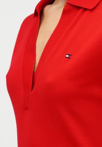 Κόκκινο πόλο με υφή υφάσματος. Διαθέτει βαθύ V λαιμό και μικρό λογότυπο σε μπλε, λευκό και κόκκινο στο αριστερό στήθος.