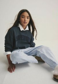 Marineblaues Sweatshirt mit "SUPERDRY" vorne gestickt, darunter ein weißes Hemd mit Kragen und helle, gestreifte, weit geschnittene Hose.