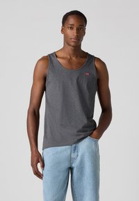 Débardeur en coton gris avec un col rond et un petit patch logo rouge, associé à un jean en denim bleu clair. Texture lisse, design décontracté.