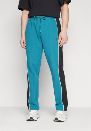 Mann trägt türkis-schwarze Trainingshose mit Seitenstreifen und weiße Turnschuhe, steht vor einem einfarbigen hellen Hintergrund.