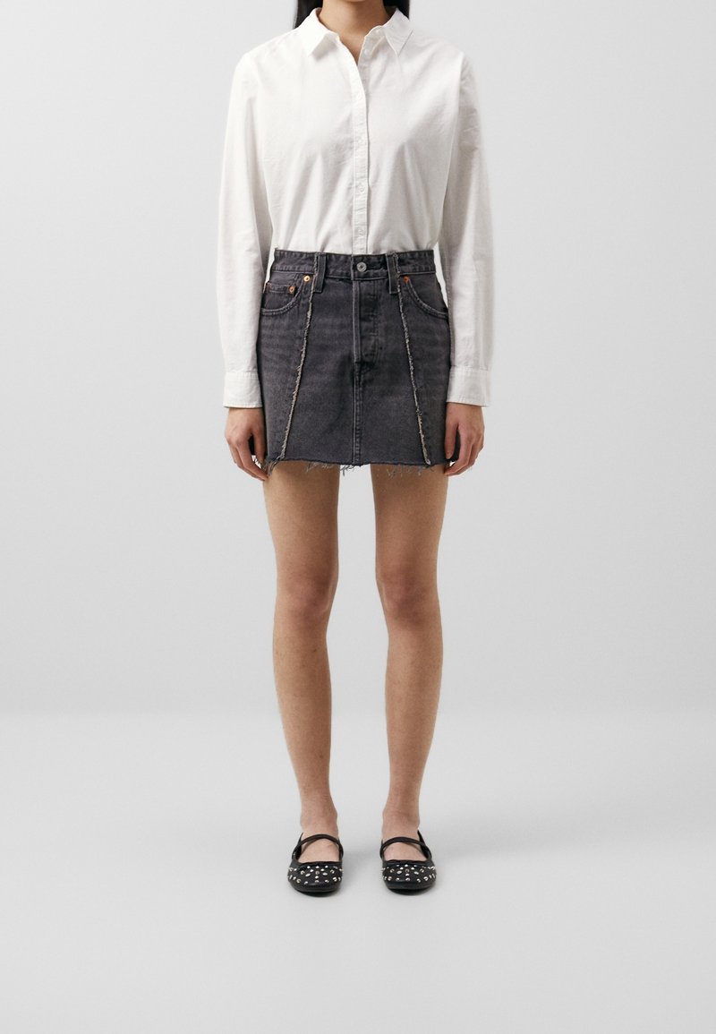 Black denim mini skirt with frayed hem, featuring striped contrast stitching and front pockets; paired with a white button-up shirt and black flats.