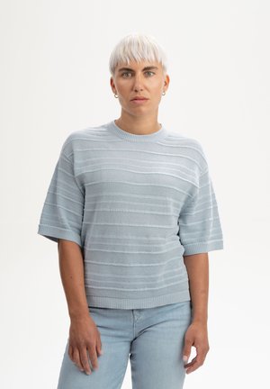 Person mit kurzen weißen Haaren, trägt einen hellblauen, kurzärmeligen Strickpullover und hellblaue Jeans, steht vor einem weißen Hintergrund.