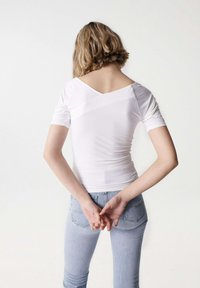 Maglietta bianca a coste con maniche corte, caratterizzata da un design con scollatura a V sulla schiena. Abbinata a jeans aderenti azzurri. Vista da dietro, mani incrociate dietro la schiena.