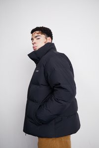 Schwarze Steppjacke mit hohem Kragen, gestepptem Muster, Seitentaschen und elastischen Bündchen. Getragen über hellen Cordhosen.