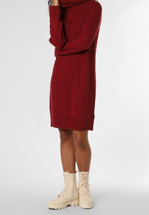 Woman wearing a red cable-knit turtleneck sweater dress with beige lace-up combat boots, standing against a plain light background.