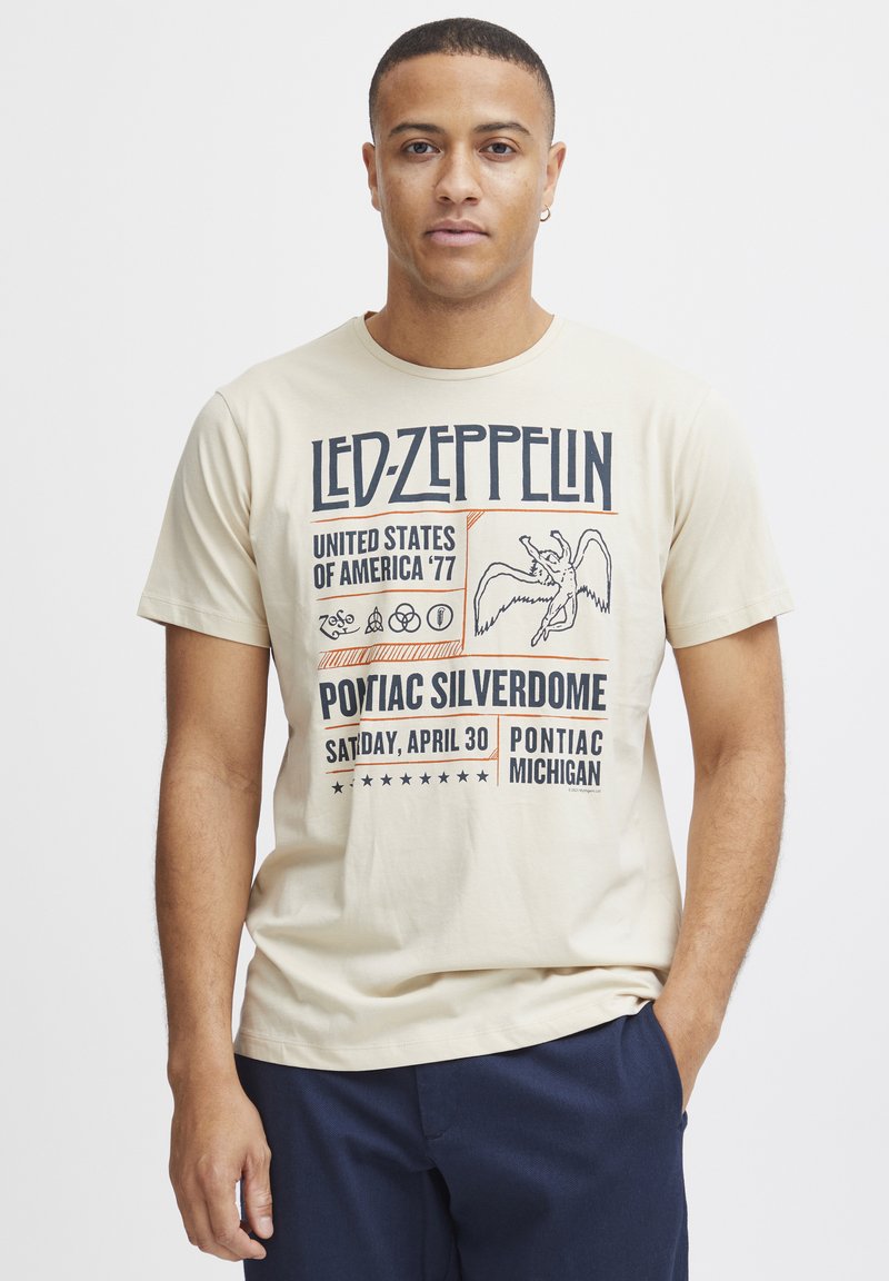 Bežs kokvilnas T-krekls, kas rotāts ar Led Zeppelin grafiku ar izteiktiem melniem un oranžiem burtiem. Dizainā ir ilustrācija ar spārnotu figūru.