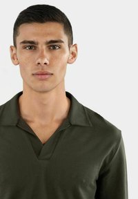 Dunkelgrünes Poloshirt mit V-Ausschnitt und kurzen Ärmeln. Der Stoff wirkt strukturiert und leicht dehnbar, mit sichtbaren Nähdetails.