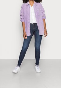Camisa de pana color lavanda, abotonada, combinada con jeans ajustados azul oscuro y zapatillas blancas. Presenta mangas arremangadas y un bolsillo en el pecho.