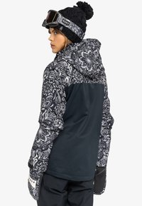 Schwarz-weißer, gemusterter Wintermantel mit einem dunklen, einfarbigen Rücken und Kapuze, kombiniert mit einer schwarzen Strickmütze und Skibrille.