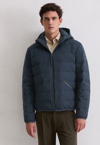 Gewatteerd jack in diep marineblauw. Glad, doorgestikt materiaal met een capuchon, een rits aan de voorkant en twee zijzakken. Gecombineerd met een lichtgekleurde blouse met knopen.