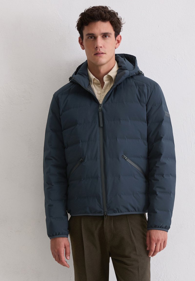 Gewatteerd jack in diep marineblauw. Glad, doorgestikt materiaal met een capuchon, een rits aan de voorkant en twee zijzakken. Gecombineerd met een lichtgekleurde blouse met knopen.