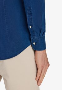 Camicia di jeans blu scuro con maniche lunghe, dotata di orlo arrotondato e polsini con bottoni. Il tessuto ha una texture liscia. Bottoni di colore chiaro visibili.