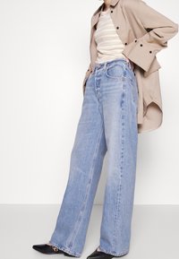 Szerokie niebieskie dżinsy z postrzępionym dołem, połączone z beżową koszulą z czarnymi guzikami i pasiastym topem bez rękawów.