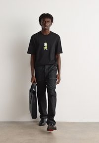 Camiseta negra con un gráfico de dinosaurio verde, combinada con jeans gris oscuro. El modelo sostiene un maletín negro y lleva zapatos negros.