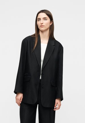 Kvinde med langt brunt hår iført en sort oversized blazer over en hvid top, stående mod en ensartet lys grå baggrund.