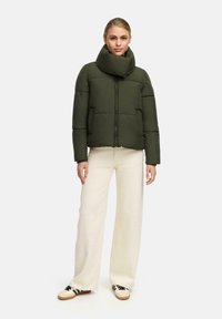 Woman stands wearing an olive green puffer jacket with high collar, wide-leg cream pants, and cream sneakers with black stripes.
