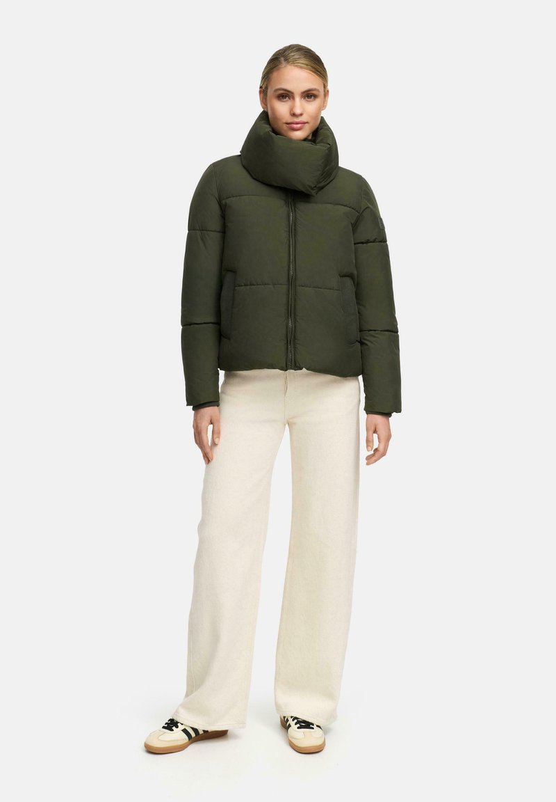 Woman stands wearing an olive green puffer jacket with high collar, wide-leg cream pants, and cream sneakers with black stripes.
