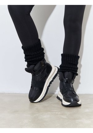 Bottes noires imperméables avec semelle blanche, dotées d'une sangle et d'un détail de fermeture éclair, associées à des chaufferettes noires côtelées sur des leggings noirs.
