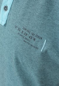 Himmelblau gestreifte Poloshirt aus Baumwolle mit einem knöpfbaren Kragen und bedruckten Textdetails, die Koordinaten und Markeninformationen enthalten.