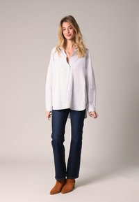 Blonde woman in loose white striped shirt, dark blue jeans, and brown ankle boots standing against a plain background, smiling.