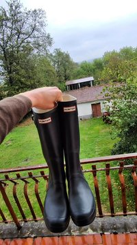 Botas de lluvia negras de goma con acabado brillante, con la marca "HUNTER" en rojo y blanco en la parte superior, textura lisa y altura media hasta la pantorrilla.