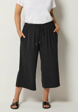 Femme portant un pantalon noir ample coupé au-dessus de la cheville avec une taille cordon et des poches latérales, associé à des sandales noires et un haut blanc à manches courtes.