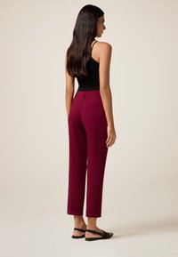 Burgunderfarbene Hosen mit maßgeschneiderter Passform, cropped Länge und umgeschlagenen Säumen, kombiniert mit einem schwarzen Tanktop und flachen Sandalen. Glatte Stofftextur.