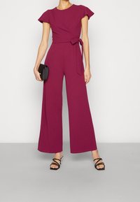 Monos vestido burdeos con diseño cruzado, mangas cortas y cinturón en la cintura. Piernas acampanadas, tejido suave, combinado con un pequeño bolso negro de mano y sandalias con tiras.