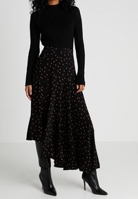Schwarzer gerippter Pullover kombiniert mit einem schwarzen Rock mit einem braunen Punktmuster. Der Rock hat einen asymmetrischen Saum. Schwarze Stiefeletten mit Absatz.