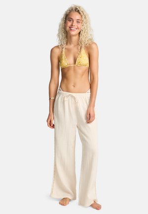 Bikini-Oberteil in gelbem Muster mit dreieckigen Cups, kombiniert mit hellbeigen, weit geschnittenen Hosen mit Seitenausschnitten und strukturierter Stoffqualität.