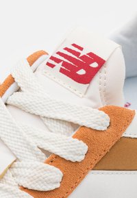 Vita sneakers med orange mockaskinaccenter, texturerade vita snören och en tygtunga med en röd logotypdesign. Slät gummisula.