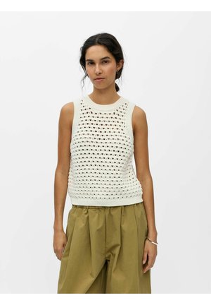 Kobieta o ciemnych włosach nosi białą, bezrękawnikową bluzkę na szydełku i luźne oliwkowo-zielone spodnie, stoi na tle jednolitego jasnego tła.
