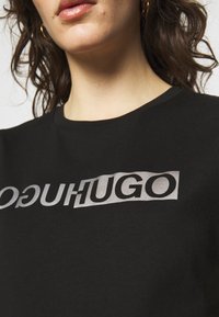 Musta puuvillainen T-paita, jossa on rohkea hopeinen logo-teksti "HUGO", klassinen pyöreä kaula-aukko ja lyhyet hihat.