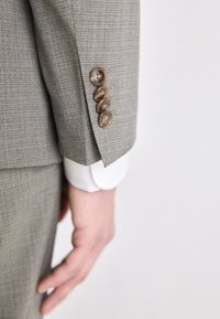 Maniche di un giubbotto grigio texturizzato con quattro bottoni marroni e il polsino di una camicia bianca sottostante. Design su misura con linee pulite e tessuto liscio.
