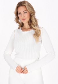 Top alb cu mâneci lungi, cu o textură nervată, guler cu margini ondulate și detalii tăiate la umeri. Designul ajustat accentuează forma.