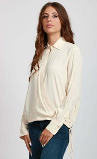 Giovane donna con lunghi capelli mossi, che indossa una blusa a maniche lunghe di colore crema con colletto e dettaglio con cravatta, abbinata a jeans blu scuro.