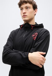 Μαύρο τζάκετ με φερμουάρ και κουκούλα, με κόκκινα κορδόνια και κόκκινο λογότυπο στο στήθος. Λείο ύφασμα και εφαρμοστό σχέδιο.