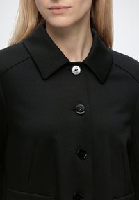 Femme aux cheveux blonds et lisses porte un manteau noir à col avec quatre boutons ; le bouton du haut est argenté, les autres sont noirs.
