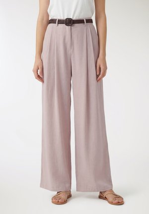 Donna che indossa pantaloni rosa chiaro a gamba larga con pieghe frontali, cintura marrone intrecciata, top bianco e sandali con cinturino intrecciato color cuoio.