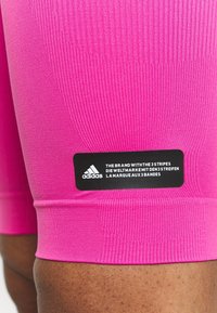 Pantaloni sportivi aderenti rosa con costine testurizzate e un'etichetta nera Adidas con testo e logo bianchi all'orlo.