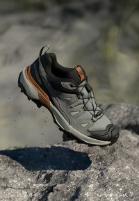 Trailrunning-Schuh für Herren, graues Mesh-Obermaterial, verstärkter schwarzer Gummikappenschuh, orangefarbene Akzente, flexible Sohle, leichtes Design, strukturierte Oberfläche.