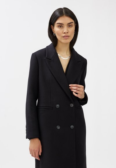 Cappotto nero doppiopetto con ampi risvolti, maniche abbottonate e tasche laterali. Realizzato in un tessuto testurizzato con una vestibilità sartoriale.