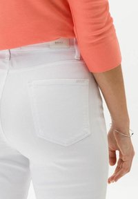 Jeans blancos con un diseño ajustado, que cuentan con un bolsillo trasero y detalles de costura sutiles. El modelo lleva una blusa color coral y una pulsera de plata.