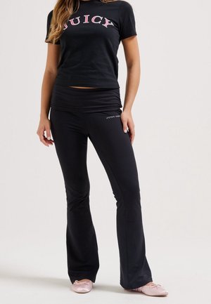 Kobieta w czarnym t-shircie Juicy Couture i dopasowanych czarnych spodniach z różowymi balerinami, stojąca na jednolitym tle.