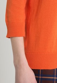 Pull orange tricoté avec un poignet côtelé, des manches trois-quarts et une coupe décontractée, associé à un pantalon à carreaux marron.