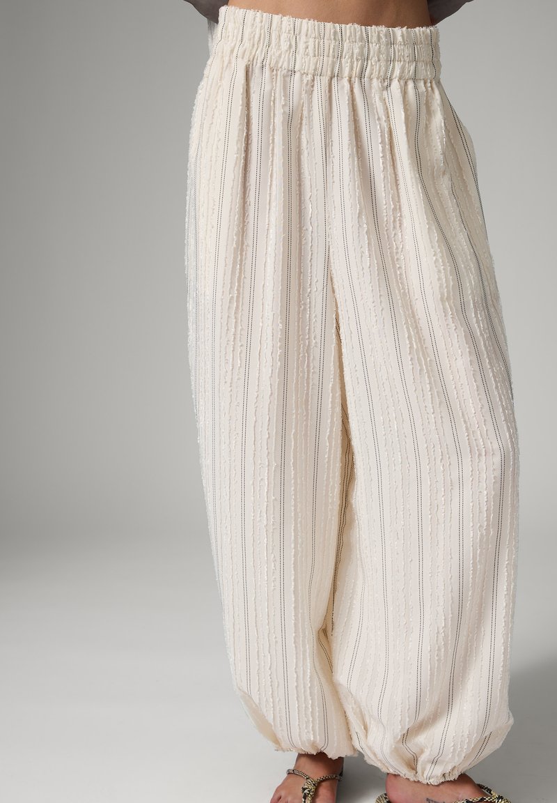 Pantalon blanc ample avec rayures texturées verticales, taille élastique et bas élastiques froncés, porté avec des sandales à bout ouvert décorées.